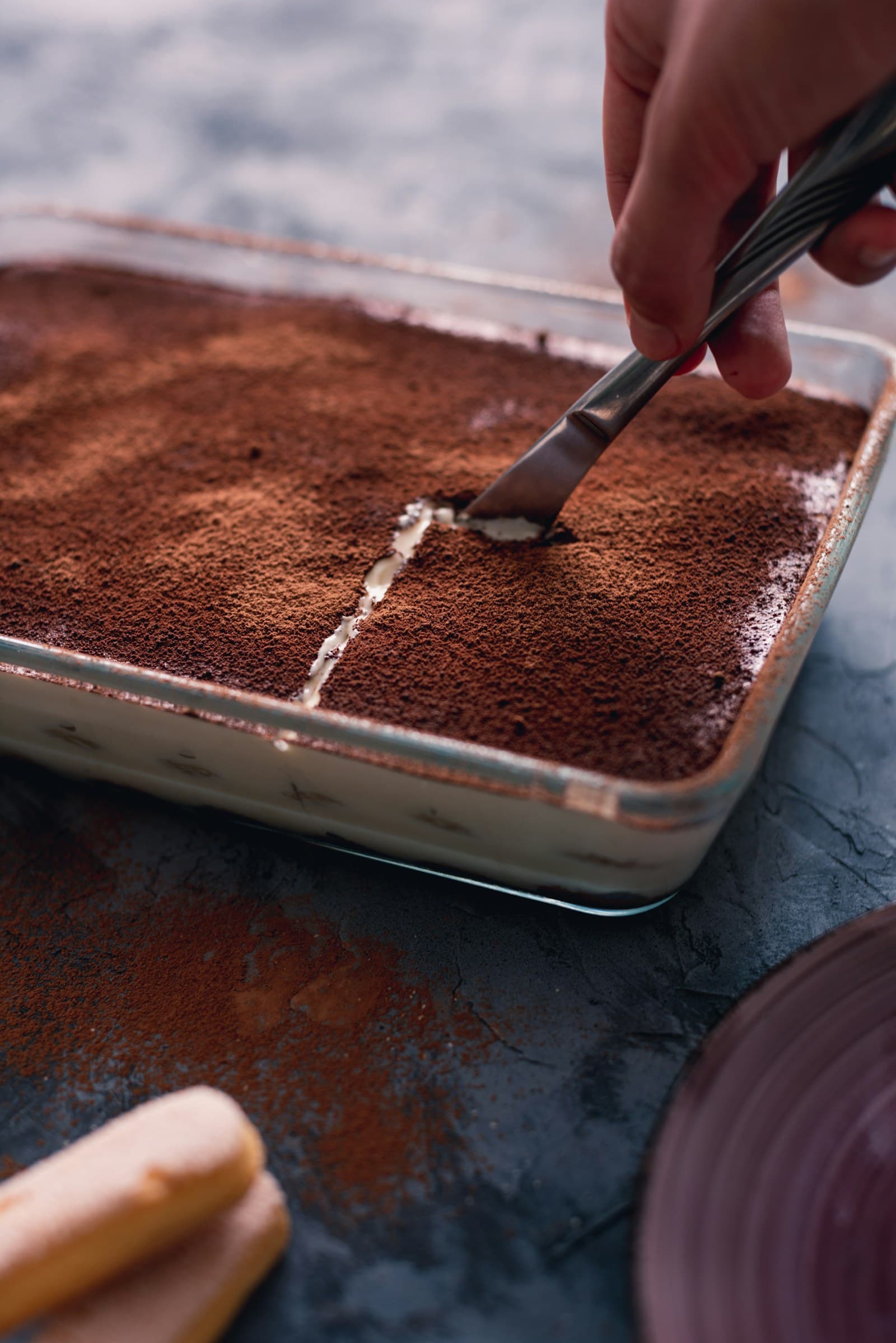 A dish of Tiramisù being cut, its cocoa surface scattered beside savoiardi biscuits